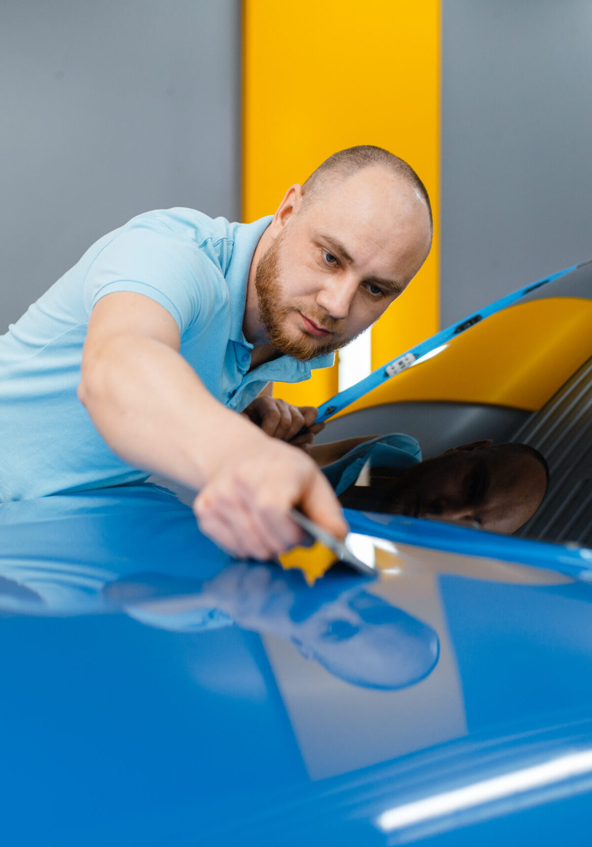 Male car wrapper with squeegee installs protective vinyl foil or film on hood. Worker makes auto detailing. Automobile paint protection coating, professional tuning